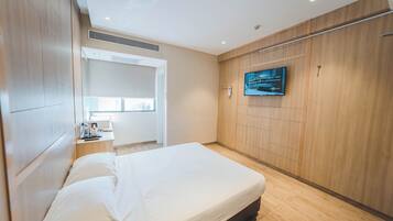 A modern hotel room with a large bed, a TV mounted on the wall, a wooden wardrobe, and a small desk with a lamp.