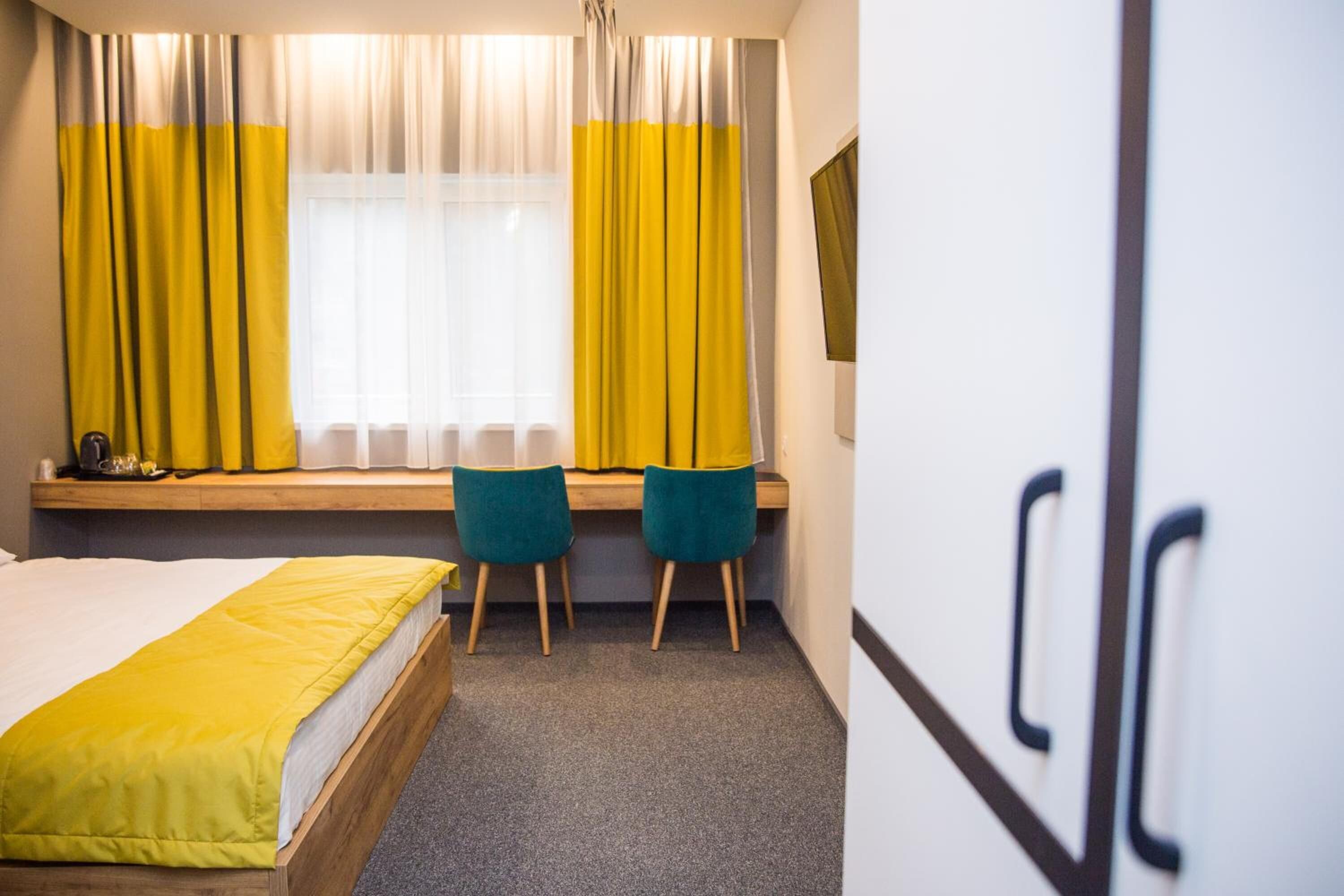 Une chambre d’hôtel avec un lit, deux chaises, une fenêtre avec des rideaux jaunes et un banc en bois.