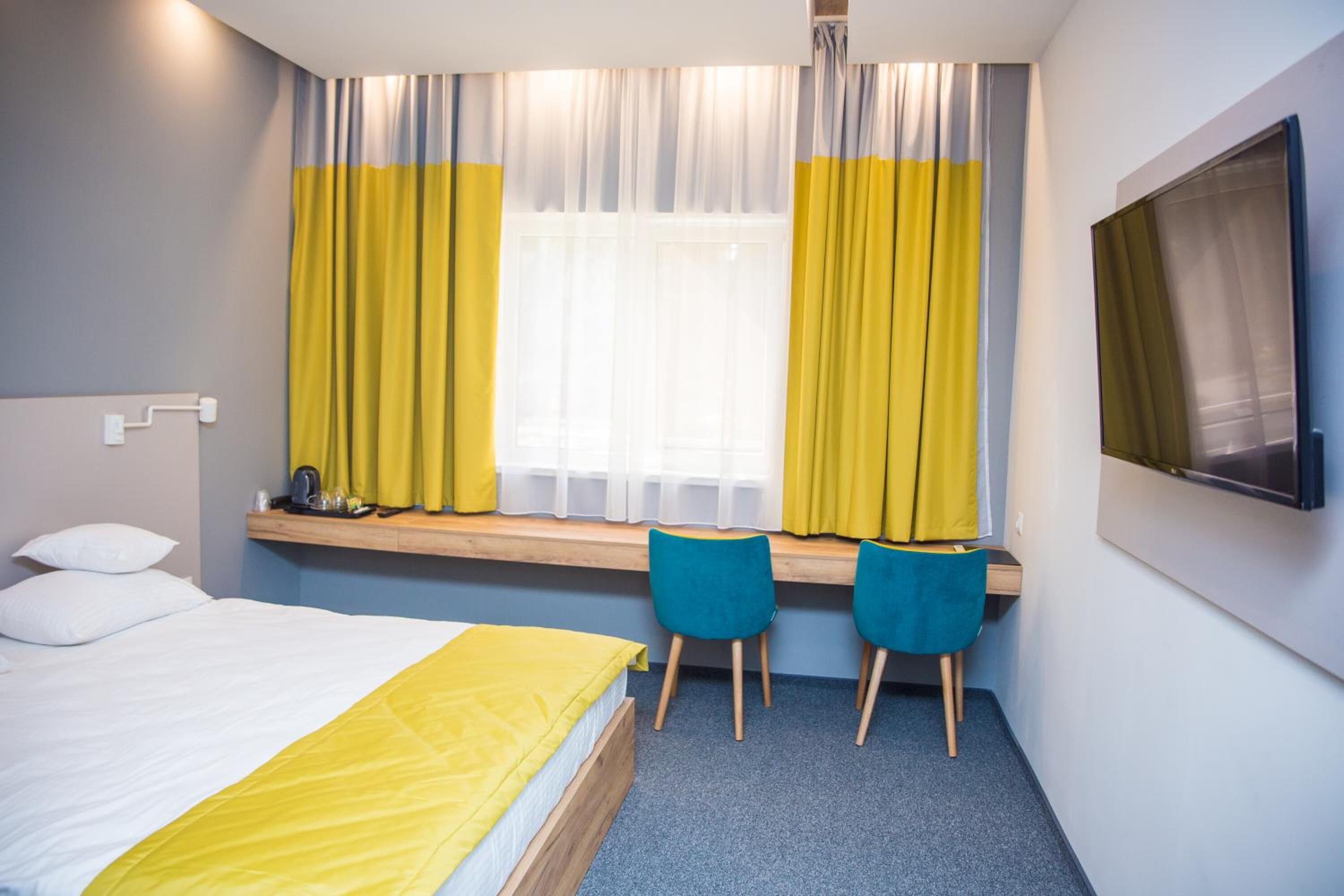 Habitación de hotel con cama, escritorio con dos sillas, ventana con cortinas y un televisor de pantalla plana montado en la pared.
