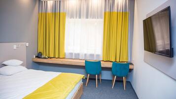Habitación de hotel con cama, escritorio con dos sillas, ventana con cortinas y un televisor de pantalla plana montado en la pared.
