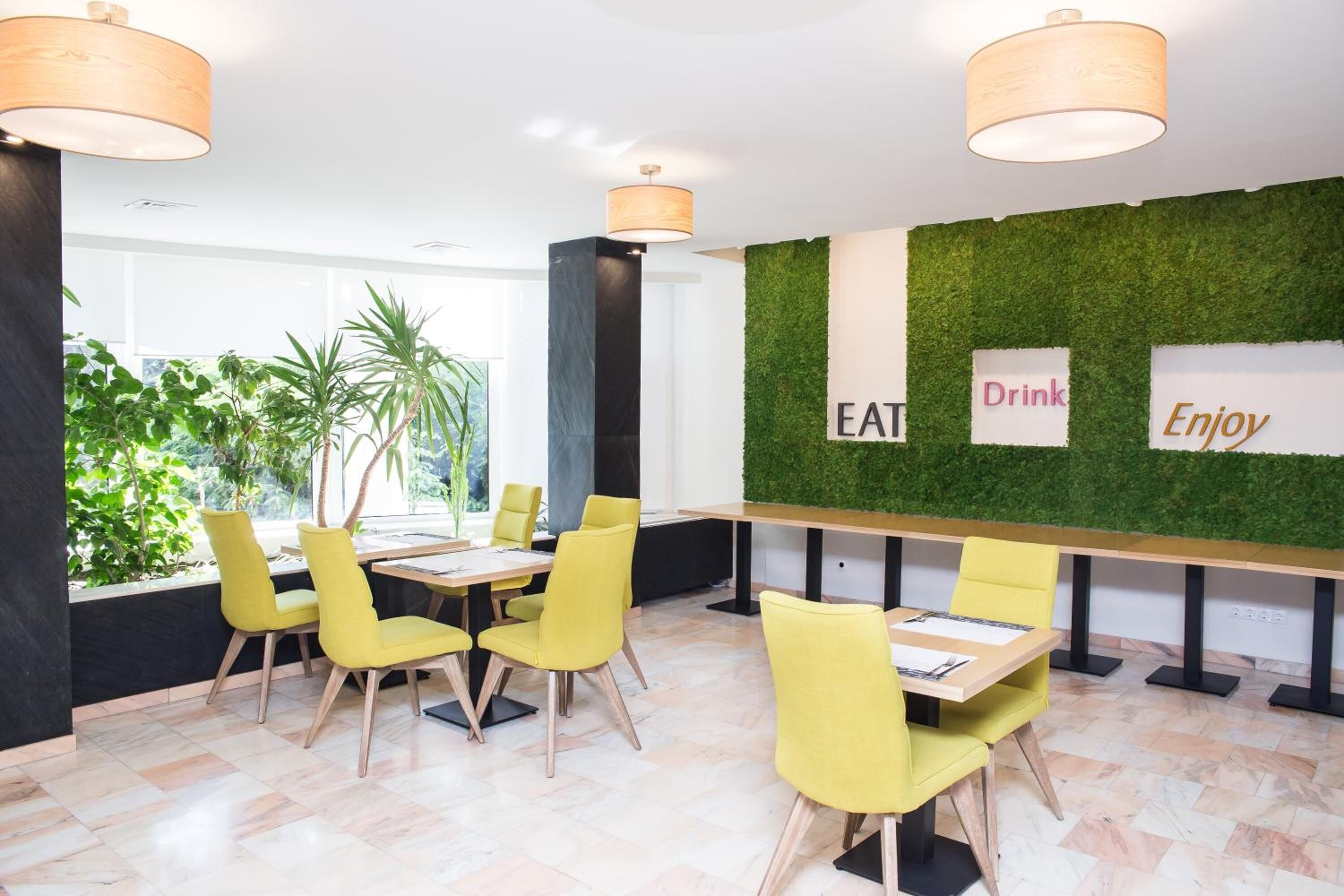 A modern dining area with yellow chairs, a long table, and a green wall with 'EAT', 'Drink', 'Enjoy' signs.