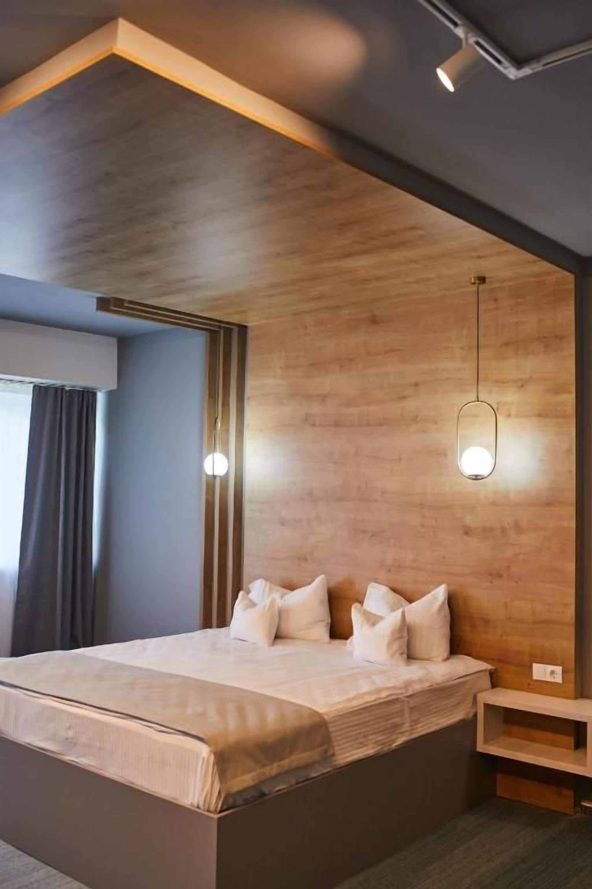 Una habitació d'hotel moderna amb un panell de fusta al capçal, un llit gran amb almoades, una taula de nit i una làmpada penjant.