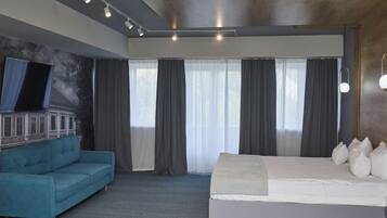 Habitació d'hotel moderna amb un llit gran, un sofà blau, una televisió de pantalla plana penjada a la paret i una finestra gran amb cortines.
