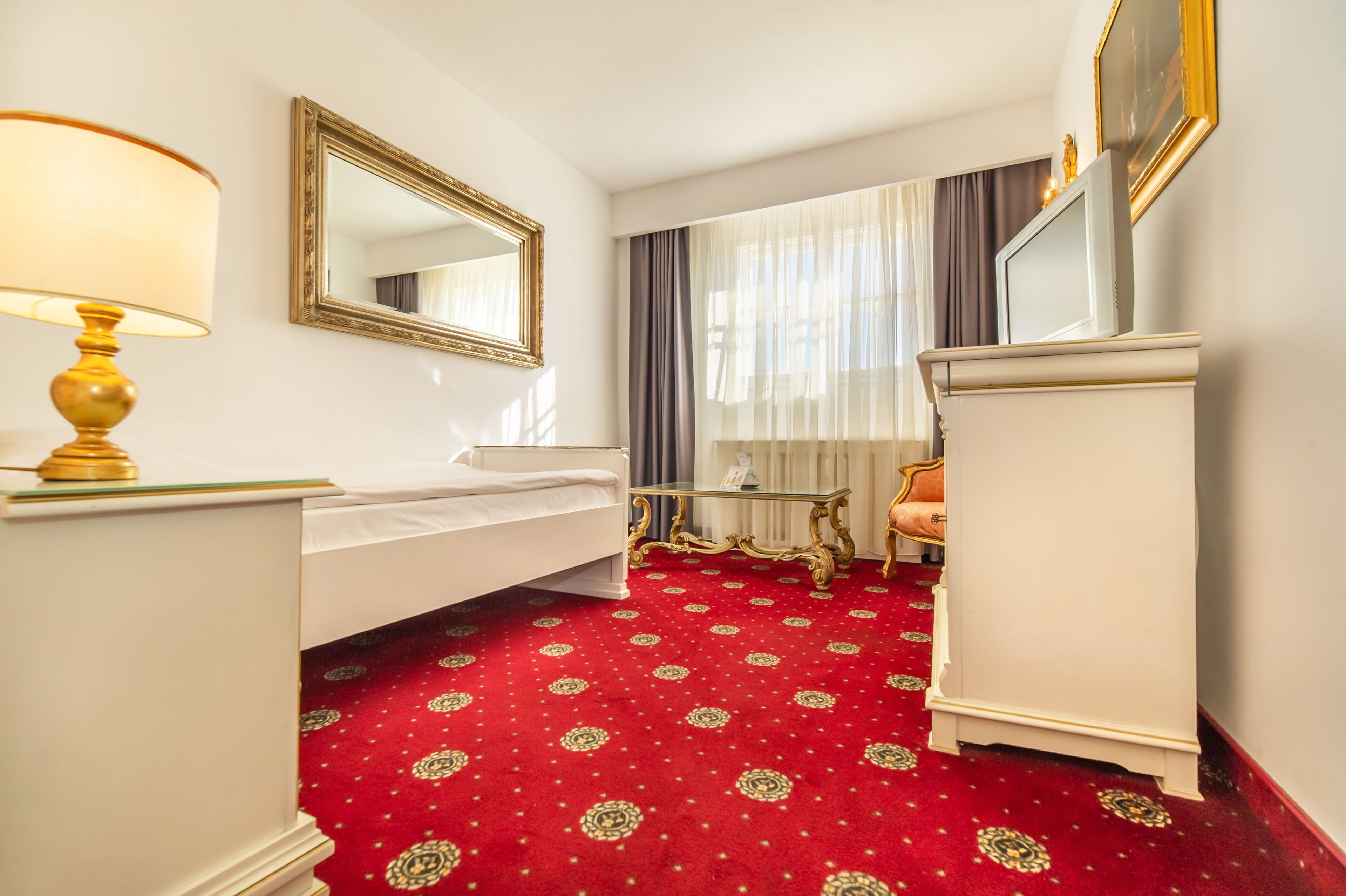 A hotel room with a red carpet, a white bed, a gold lamp, a mirror, a dresser, and a window with curtains.