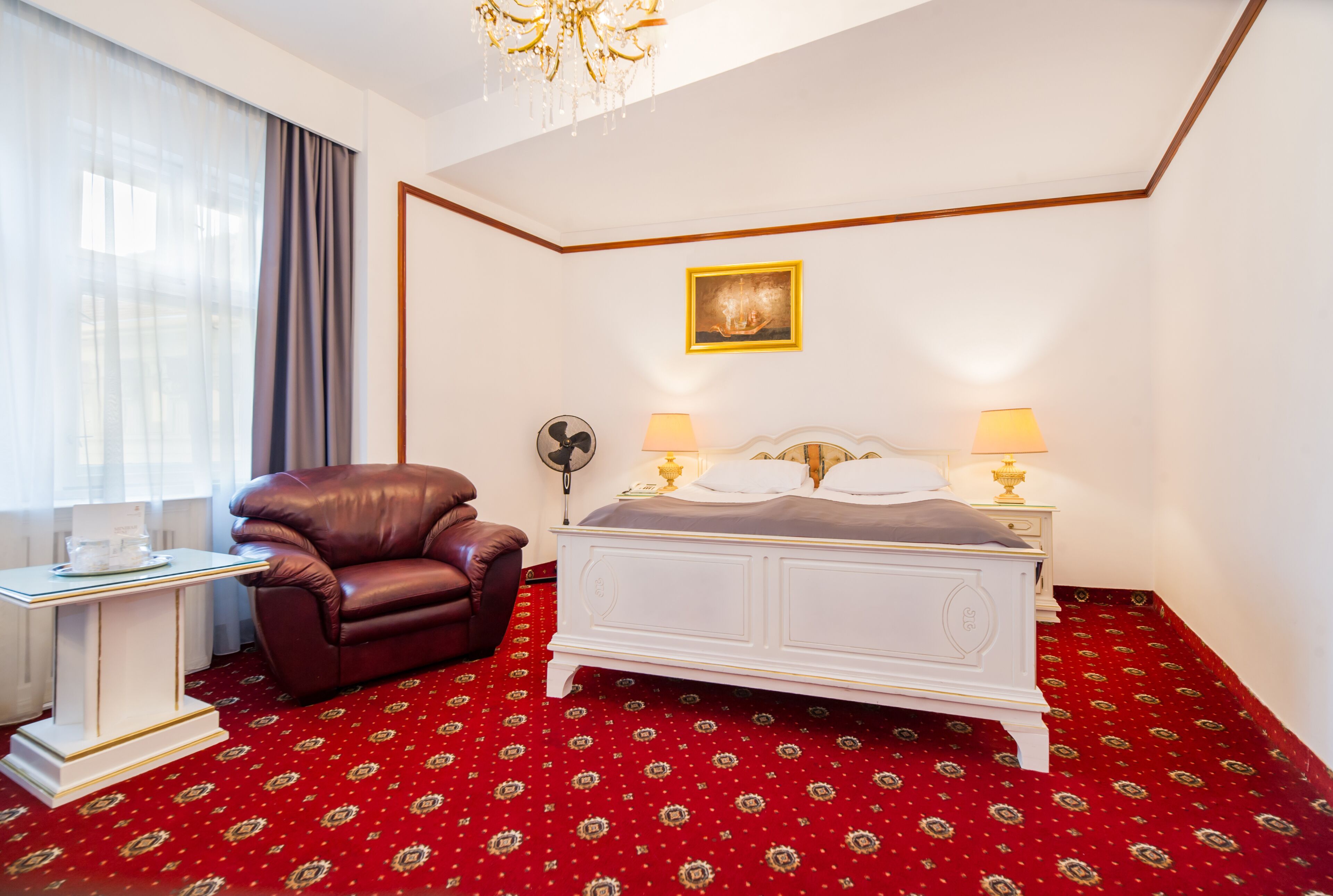 A hotel room with a large bed, a leather armchair, a bedside table, and a chandelier.