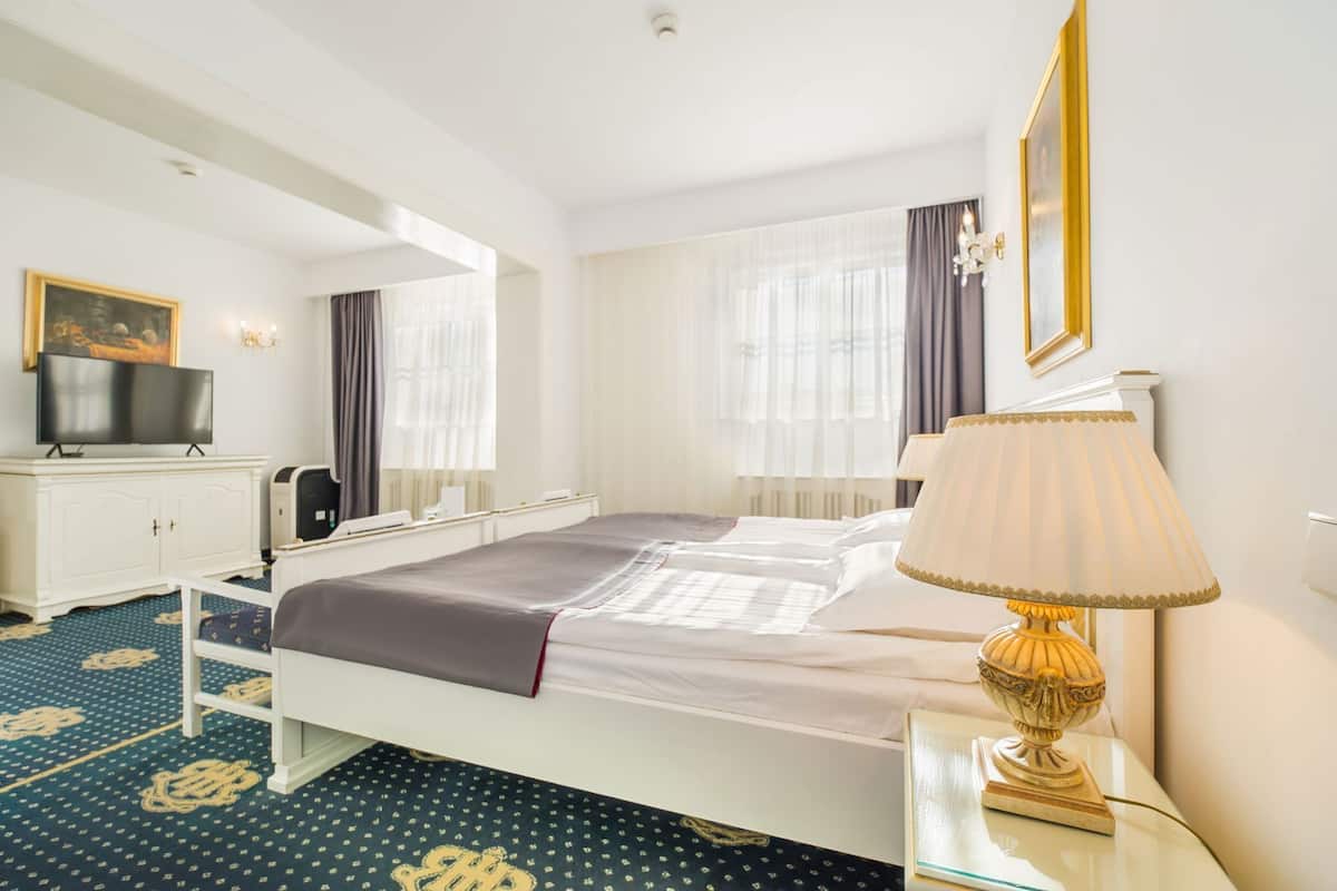 A hotel room with a bed, a desk with a lamp, a television, and a large window with curtains.