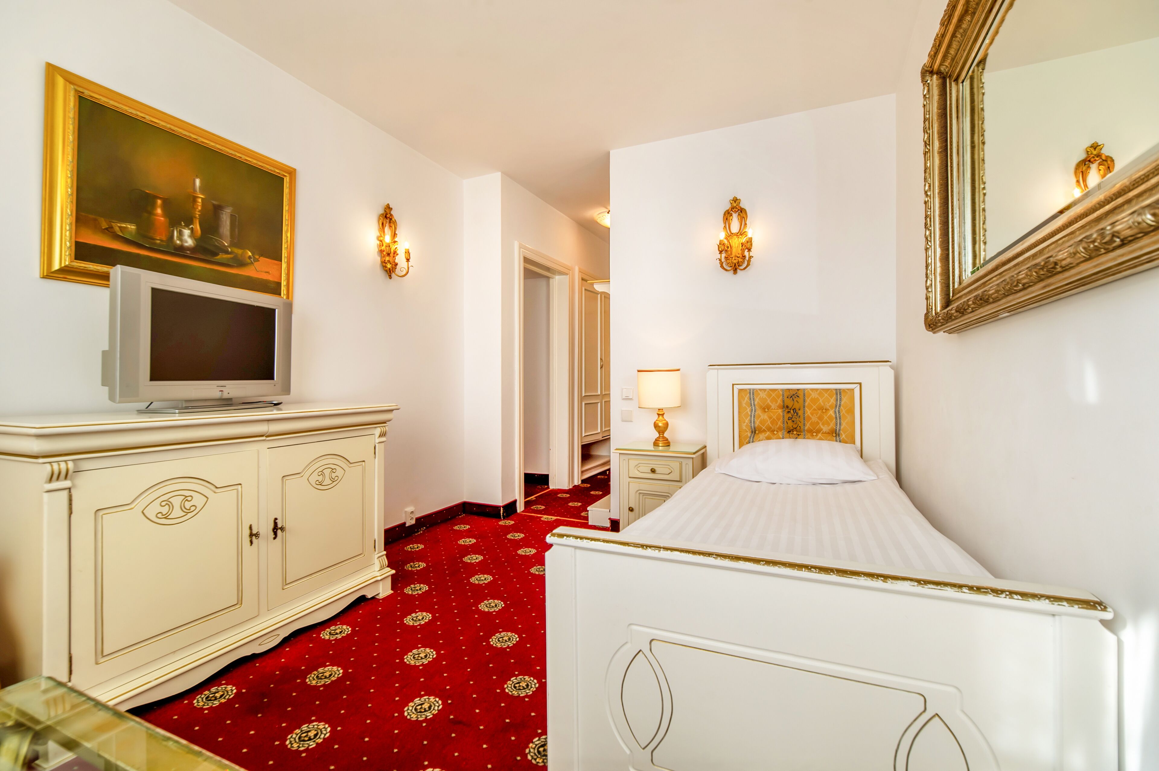 A hotel room with a bed, a television on a cabinet, a painting on the wall, and a red carpet.