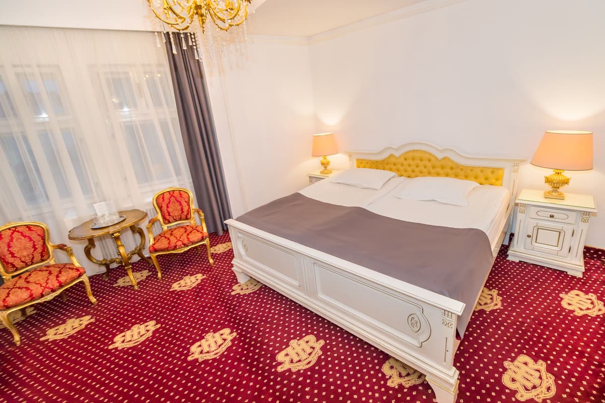A hotel room with a large bed, two bedside tables with lamps, a small round table, two armchairs, and a chandelier.