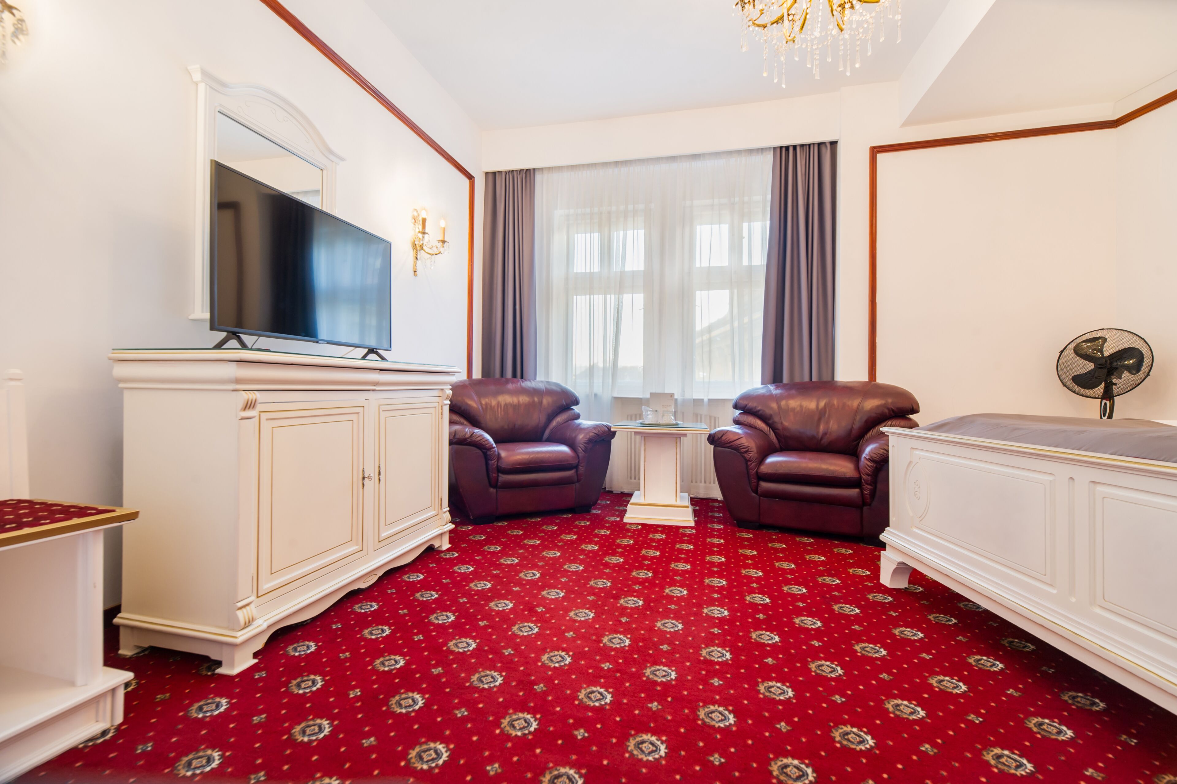 A room with a red patterned carpet, a television on a white cabinet, two leather chairs, a small table, and a window with curtains.