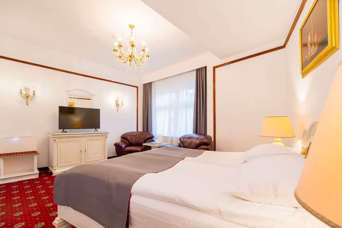 A hotel room with a large bed, a TV, leather chairs, a desk, and a chandelier.
