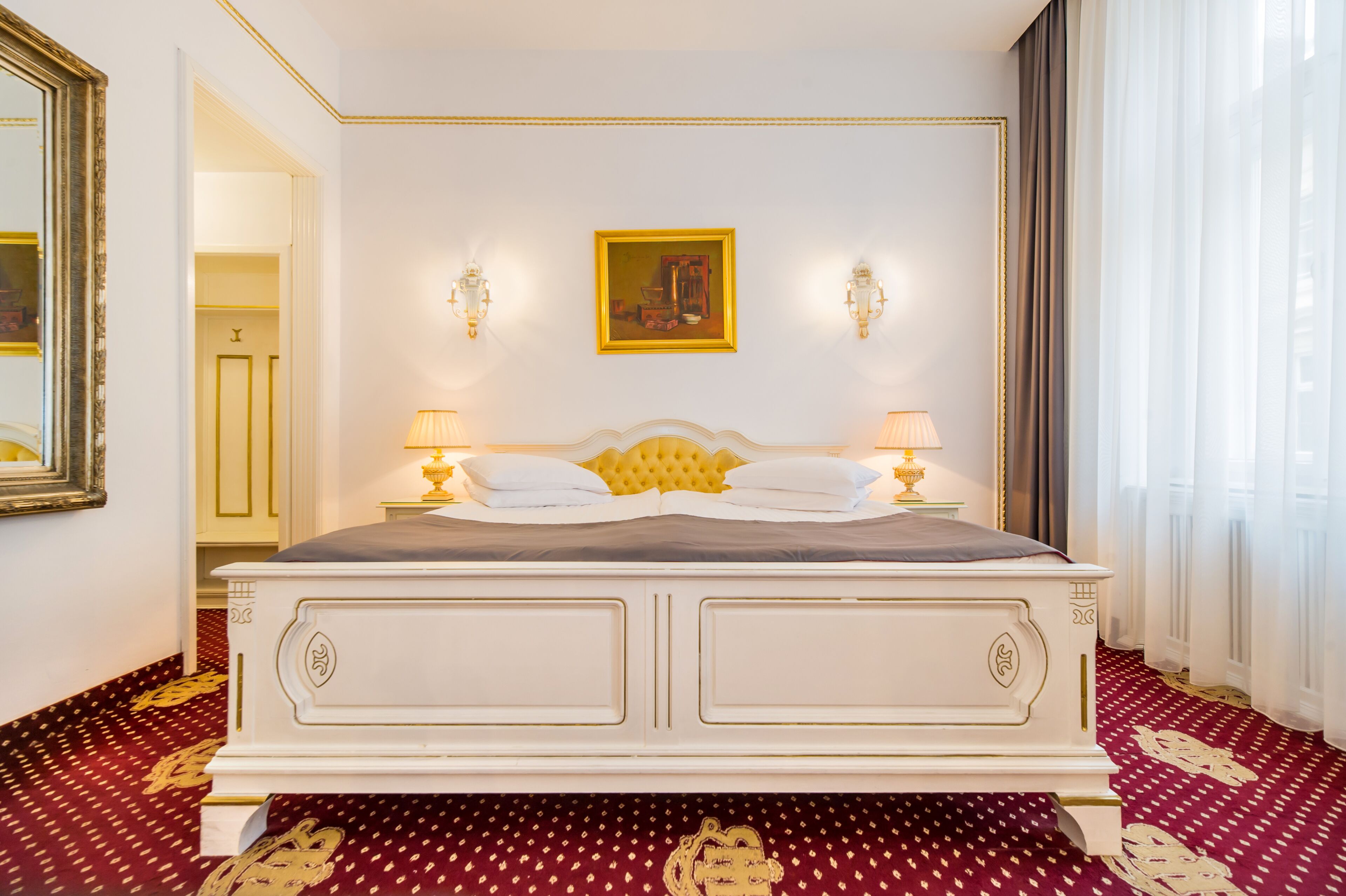 A large bed with a white frame and gold accents, two wall-mounted lamps, a framed painting above the bed, and a red patterned carpet.