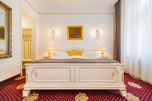 A large bed with a white frame and gold accents, two wall-mounted lamps, a framed painting above the bed, and a red patterned carpet.