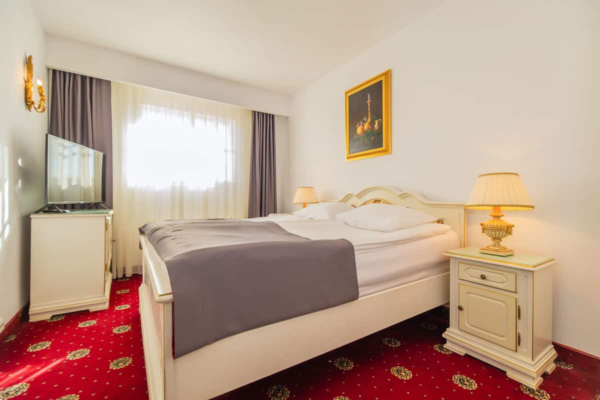 A hotel room with a bed, a bedside table with a lamp, a TV, and a painting on the wall.