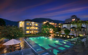Un hotel de varios pisos con piscina, sombrillas exteriores y montañas al fondo.
