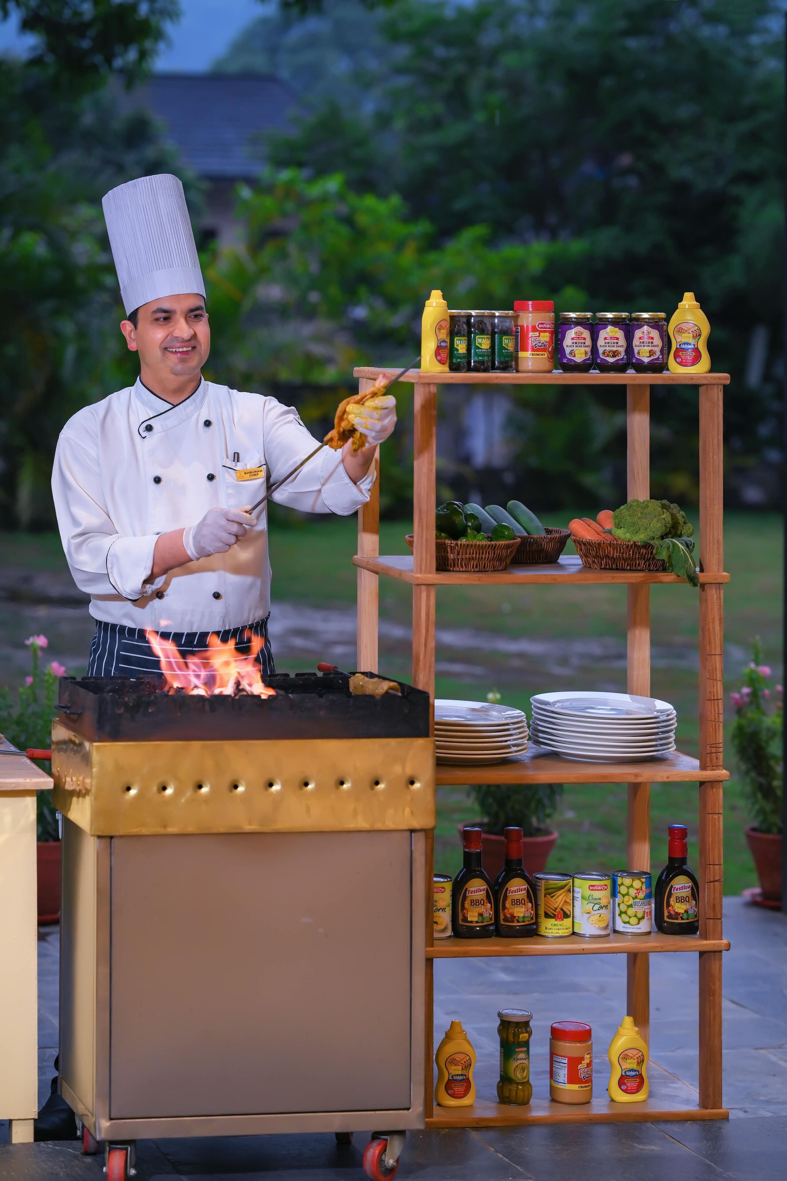 A chef grilling outdoors with a cart containing cooking equipment and condiments.