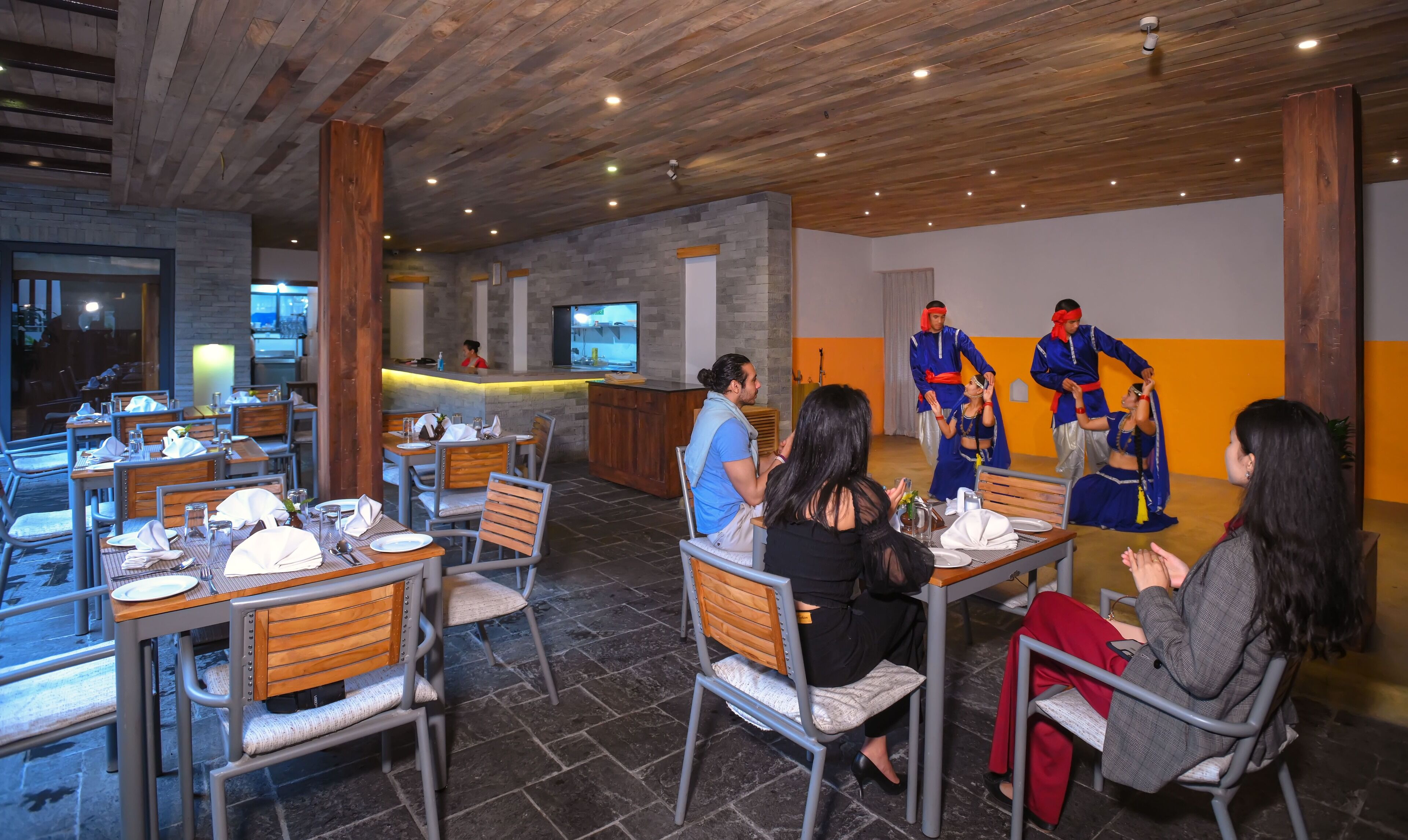 A restaurant with wooden ceiling beams, stone walls, and a counter with a TV. Tables are set with white tablecloths and chairs.
