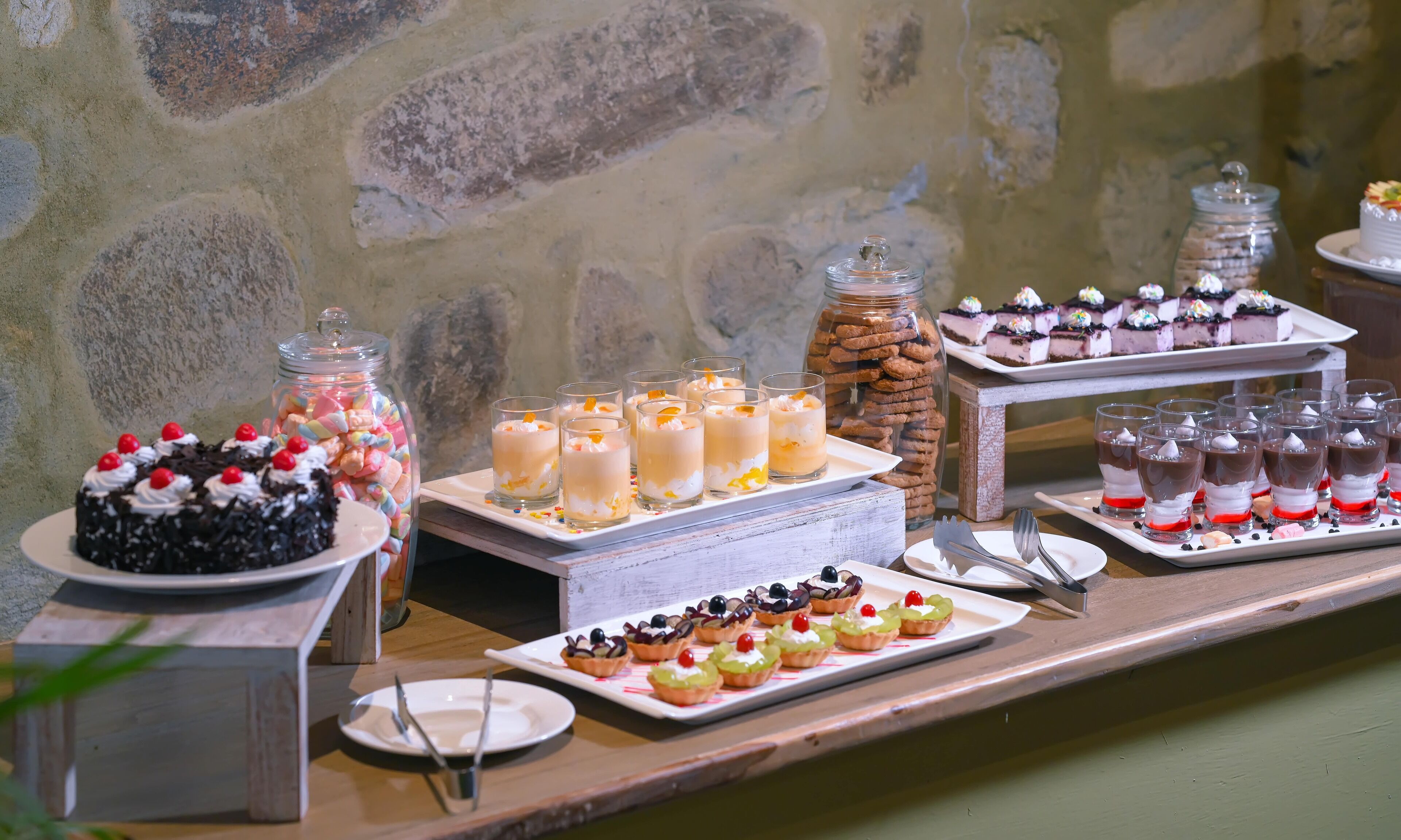 A buffet with various desserts, including cakes, cupcakes, and fruit tarts, arranged on a wooden table against a stone wall.