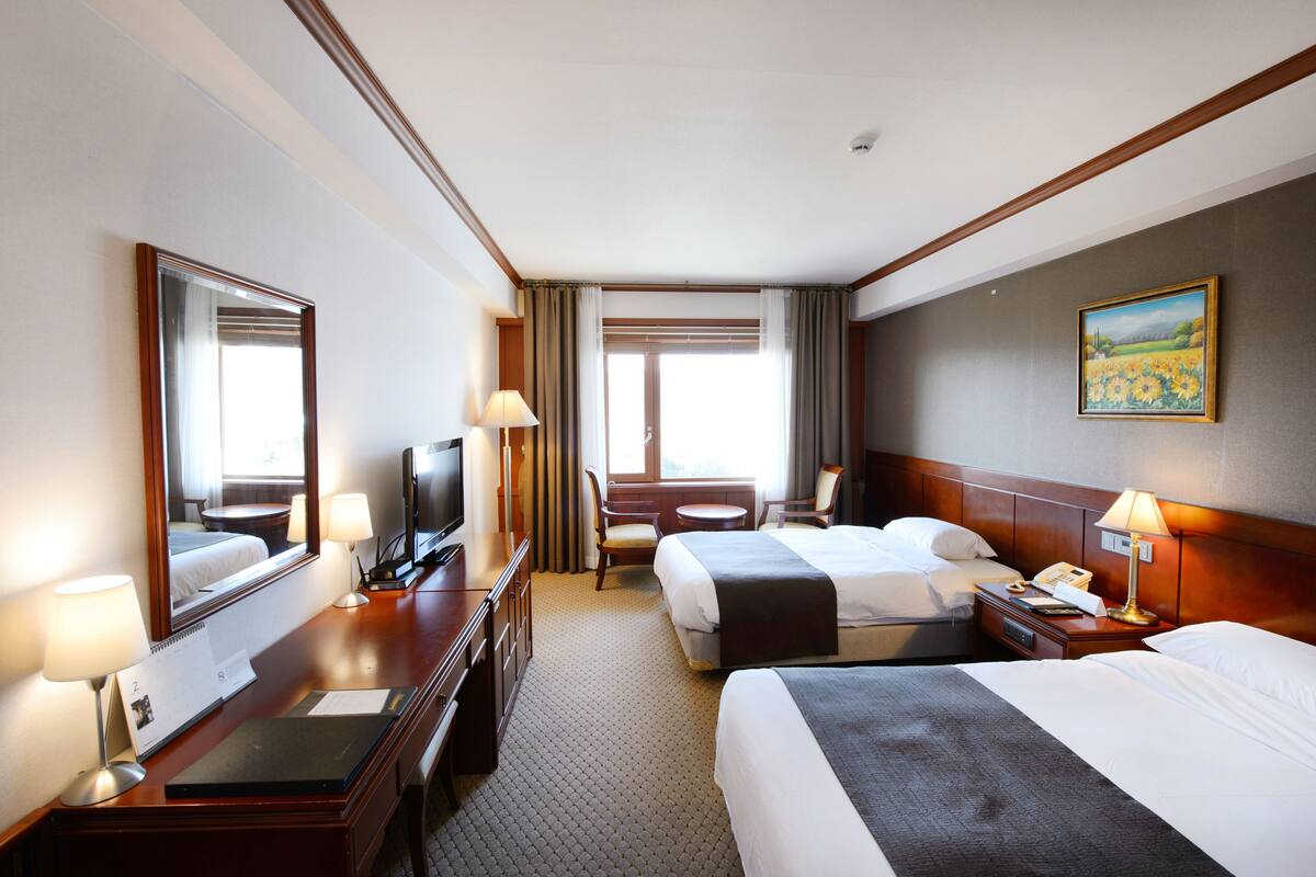 A hotel room with two beds, a desk with a TV, lamps, a painting on the wall, and a window with curtains.