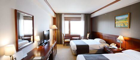 A hotel room with two beds, a desk with a TV, lamps, a painting on the wall, and a window with curtains.