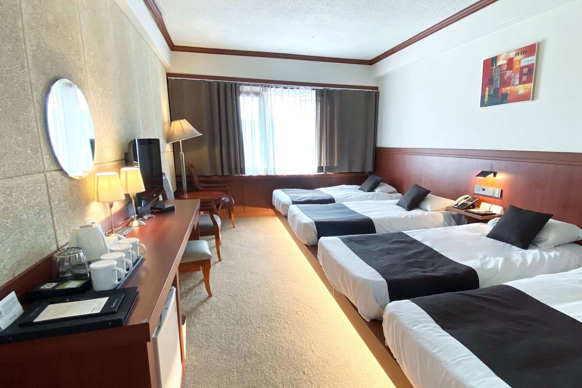 A hotel room with three beds, a desk with a computer, a chair, a lamp, and a window with curtains.