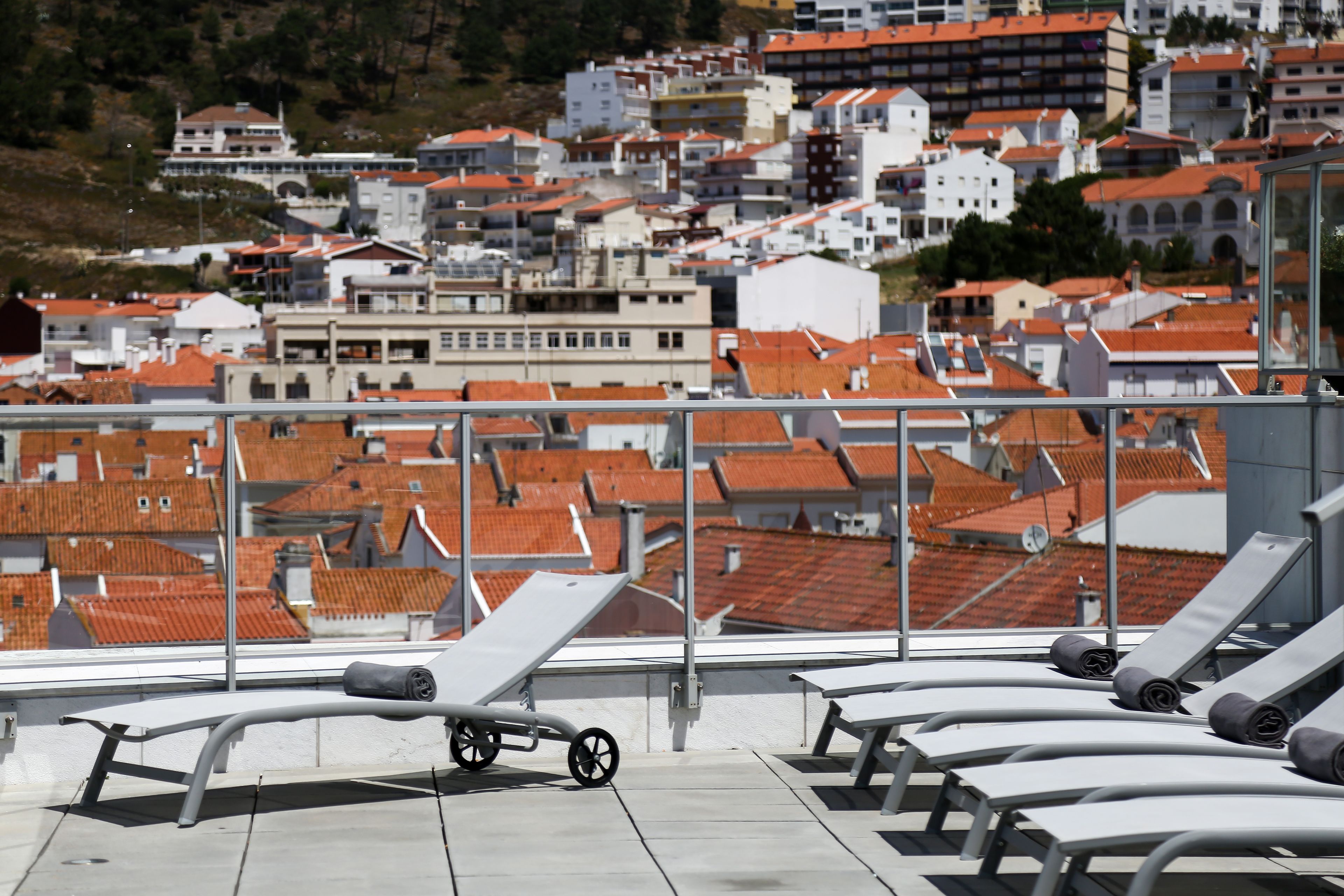 A rooftop terrace with sun loungers and a wheelchair, overlooking a densely packed urban area with red-tiled roofs.