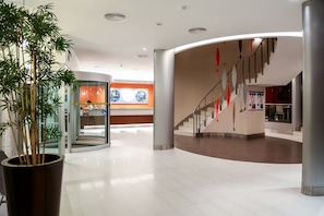 A spacious lobby with a modern design, featuring a large potted plant, a glass elevator, and a staircase with red accents. - Hotel Praia (Nazaré)
