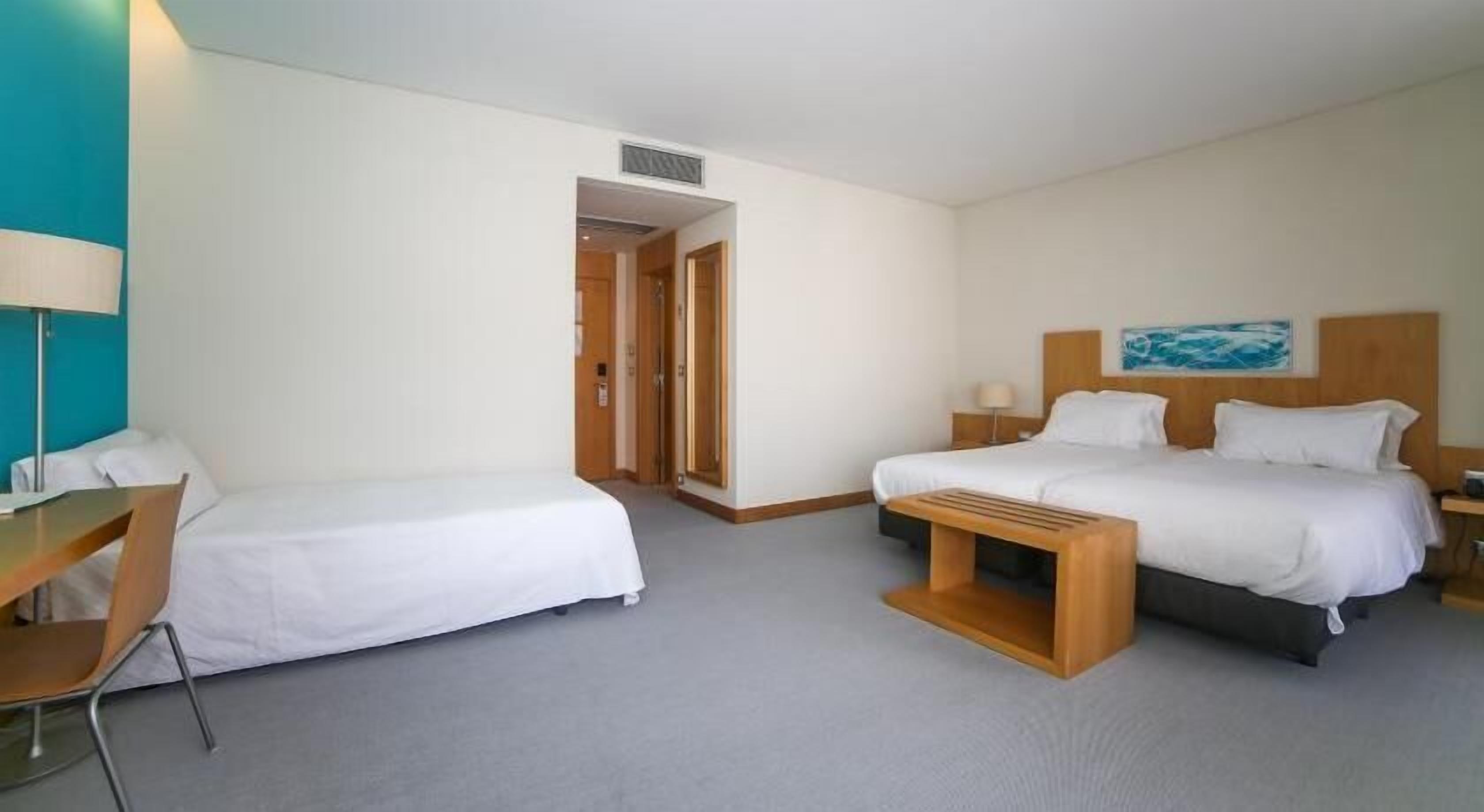 Habitación de hotel con dos camas, un escritorio y una silla. Hay un cuadro en la pared y una puerta que conduce a otra habitación.