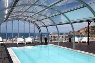 Una piscina cubierta con paredes de vidrio que ofrece vistas al mar y una terraza de madera con sillones de descanso.