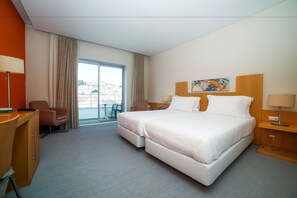 A hotel room with a large bed, a desk, a chair, and a view of the city through the window. - Hotel Praia (Nazaré)