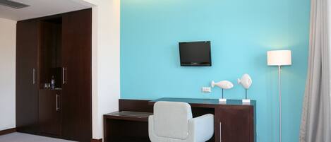 A hotel room with a teal accent wall, a dark wood desk with a flat-screen TV, a white chair, a floor lamp, and a wardrobe.