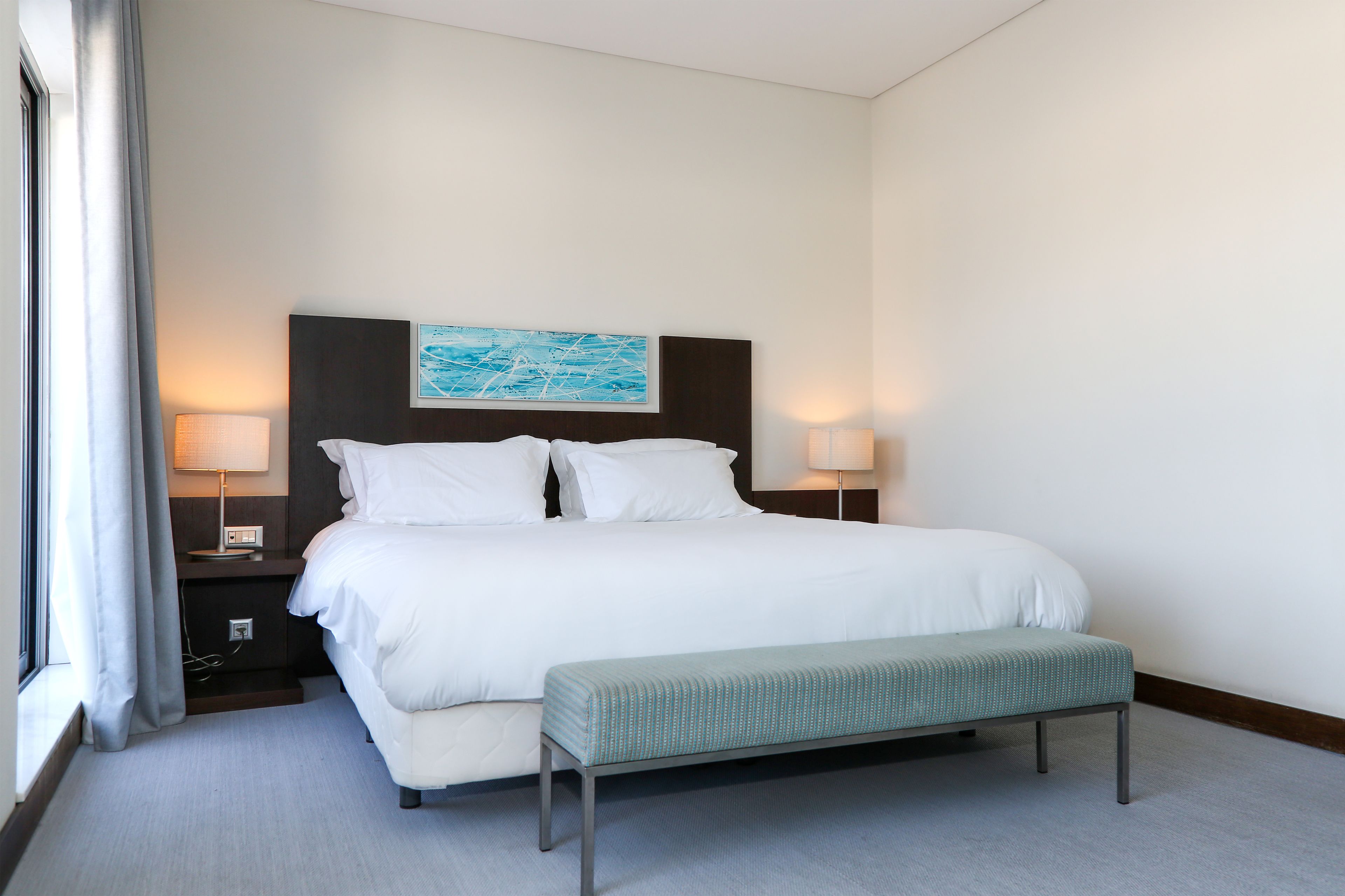 A hotel room with a large bed, two bedside lamps, a bench, and a painting on the headboard.
