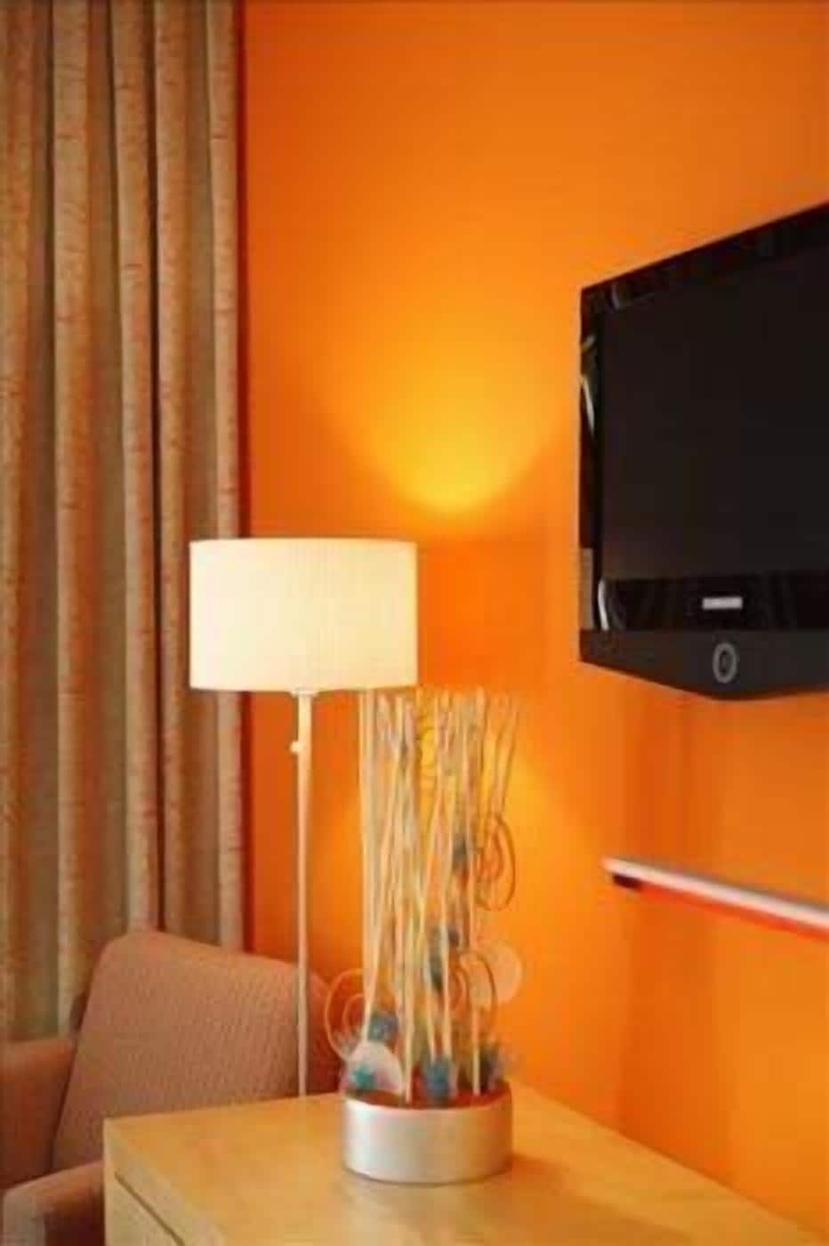 Een kamer met een oranje wand, een flatscreen televisie boven een houten tafel en een lamp met een witte kap op de tafel.