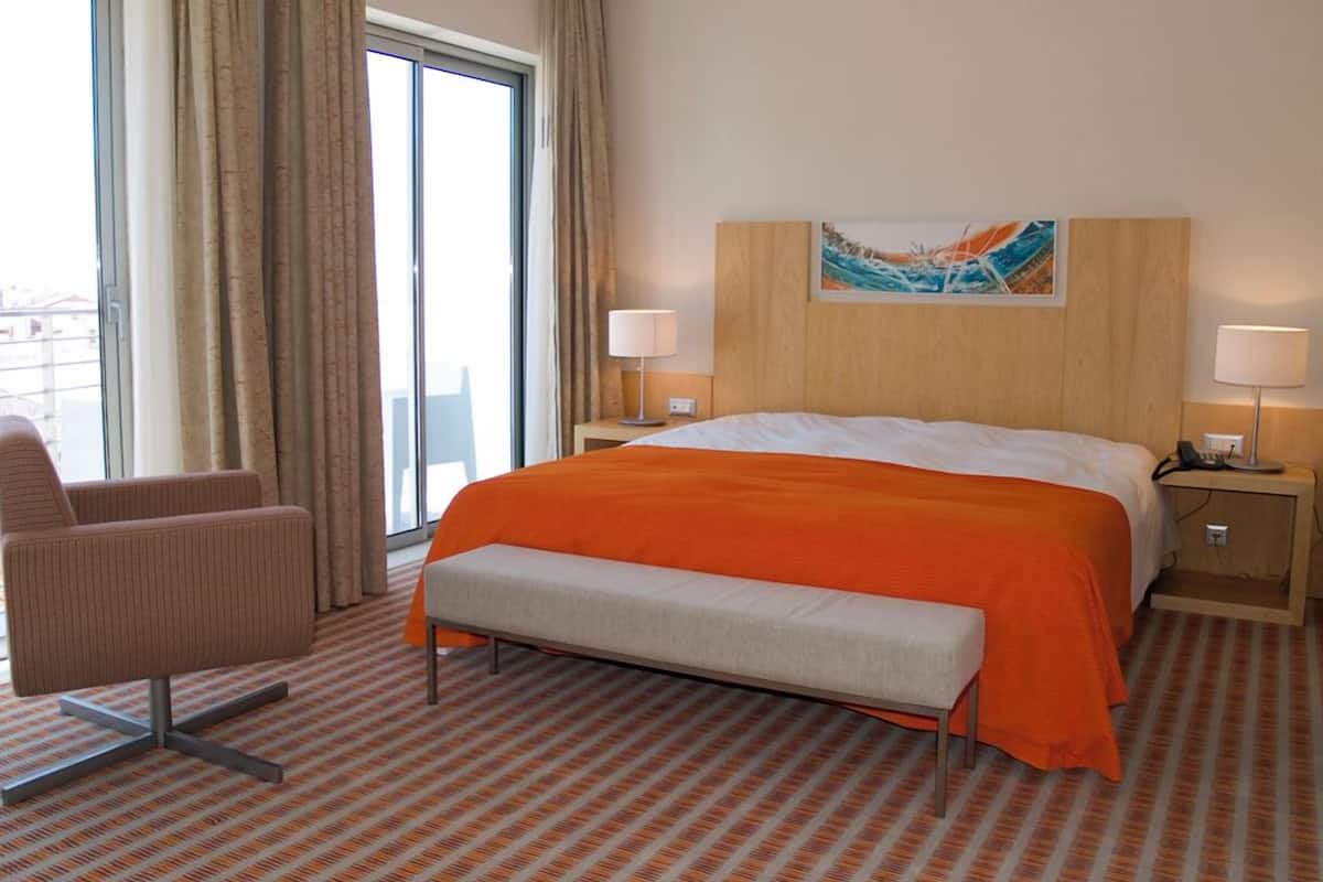 Ein Hotelzimmer mit einem großen Bett, einer Bank, einem Sessel, zwei Nachttischen mit Lampen, einem Bild an der Wand und einem Fenster mit Vorhängen.