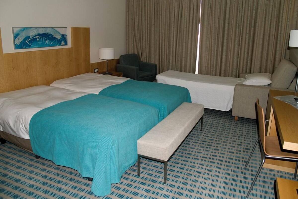 Ein Hotelzimmer mit zwei Betten, einem Schreibtisch, einem Stuhl und einem großen Fenster mit Vorhängen.