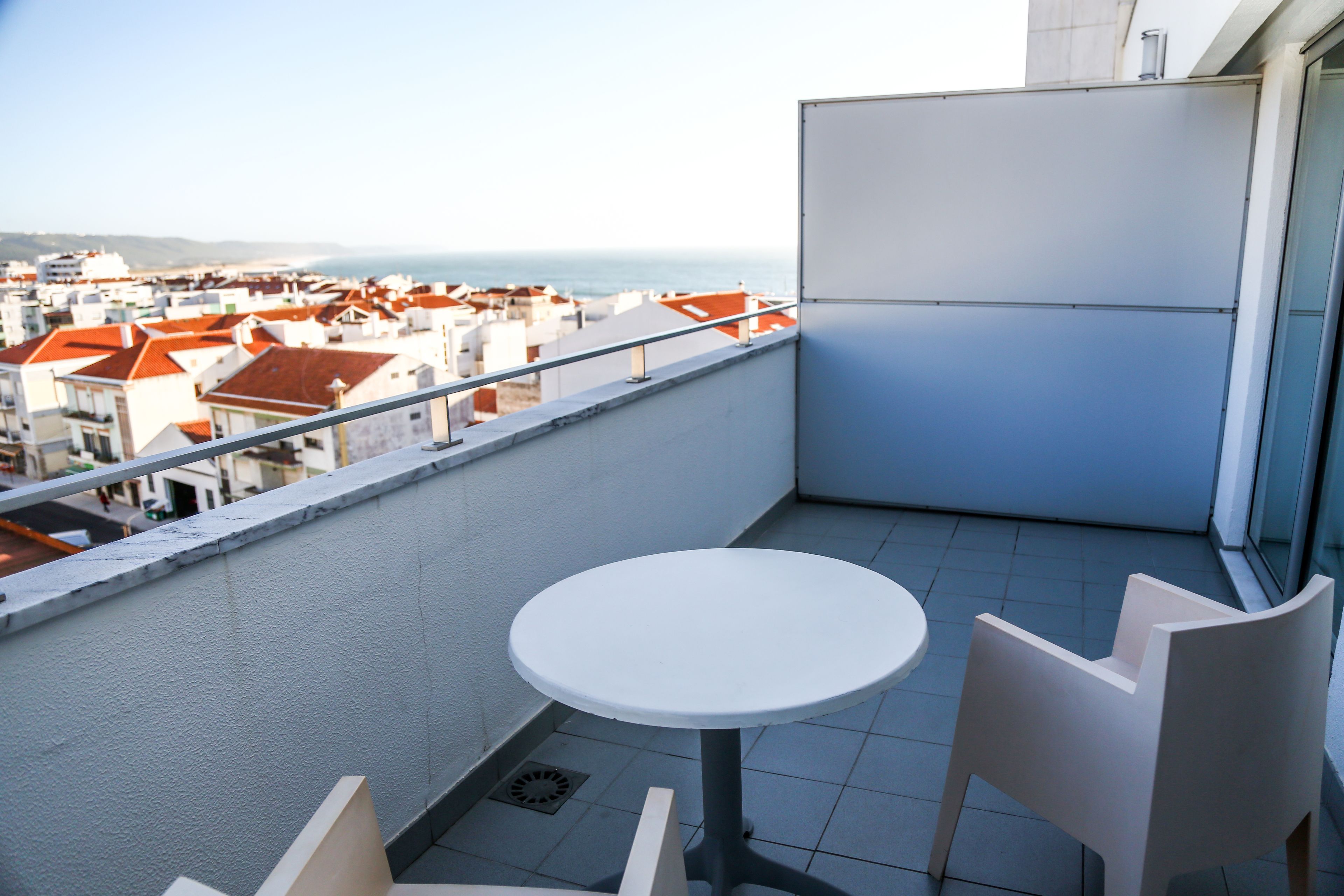 A balcony with a round table and two chairs, overlooking a cityscape with red-roofed buildings and a view of the sea.