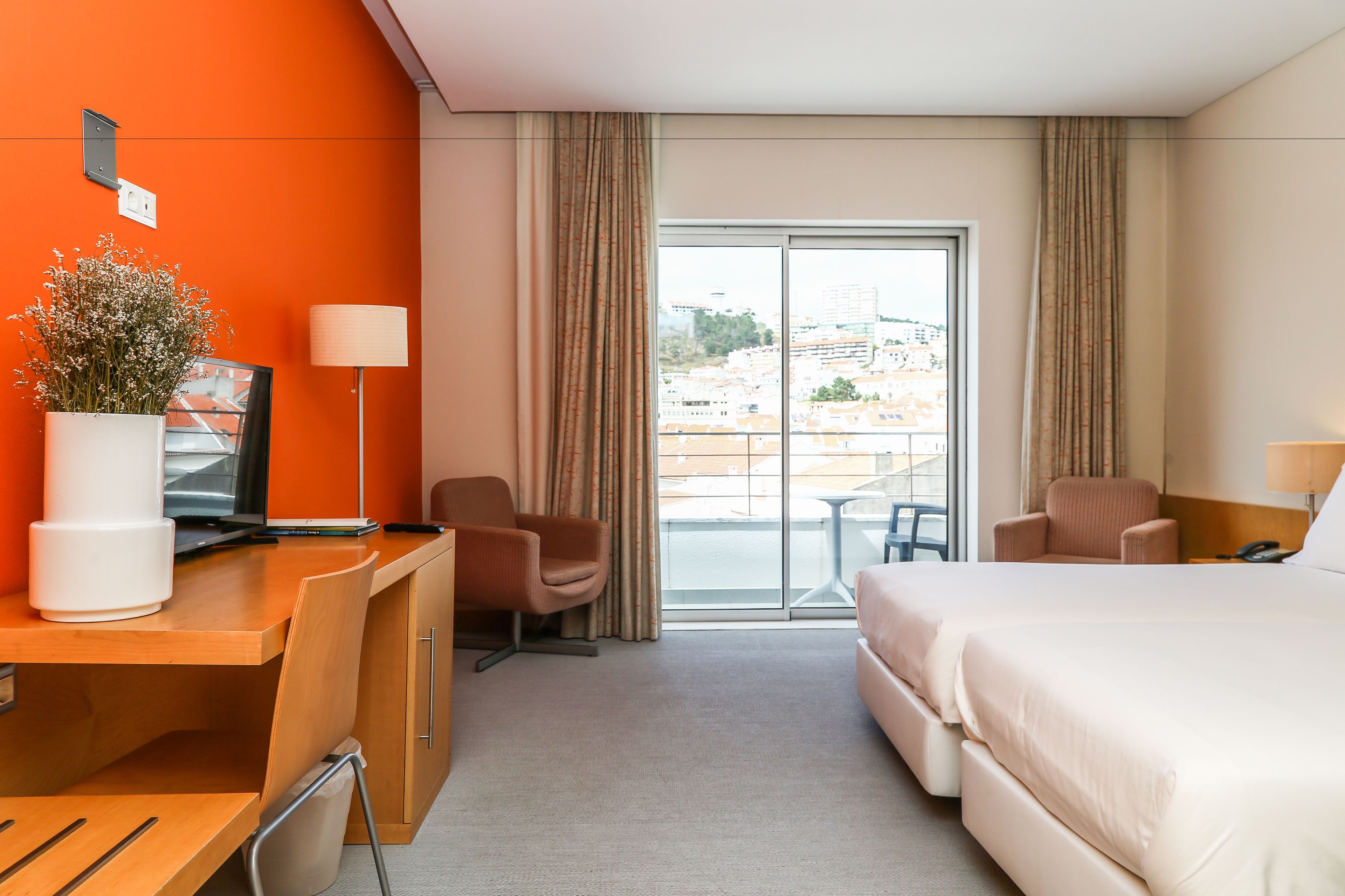 Camera d'albergo con un letto, una scrivania, sedie, una lampada, una pianta in vaso e vista sulla città dalla finestra.