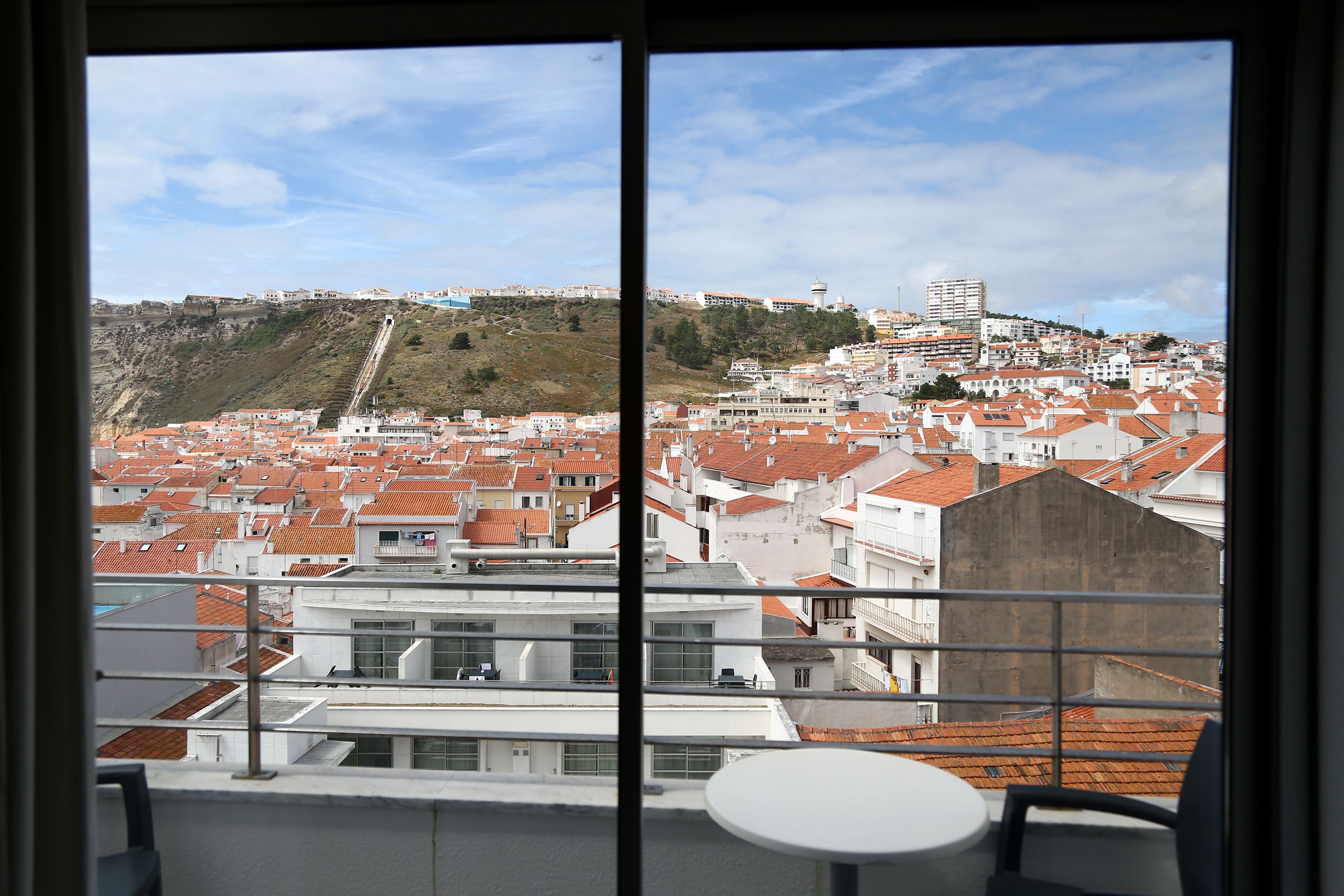 Une vue depuis un balcon donnant sur un paysage urbain, avec des bâtiments aux toits de tuiles rouges, une colline avec un escalier et un bâtiment moderne doté d’une tour.