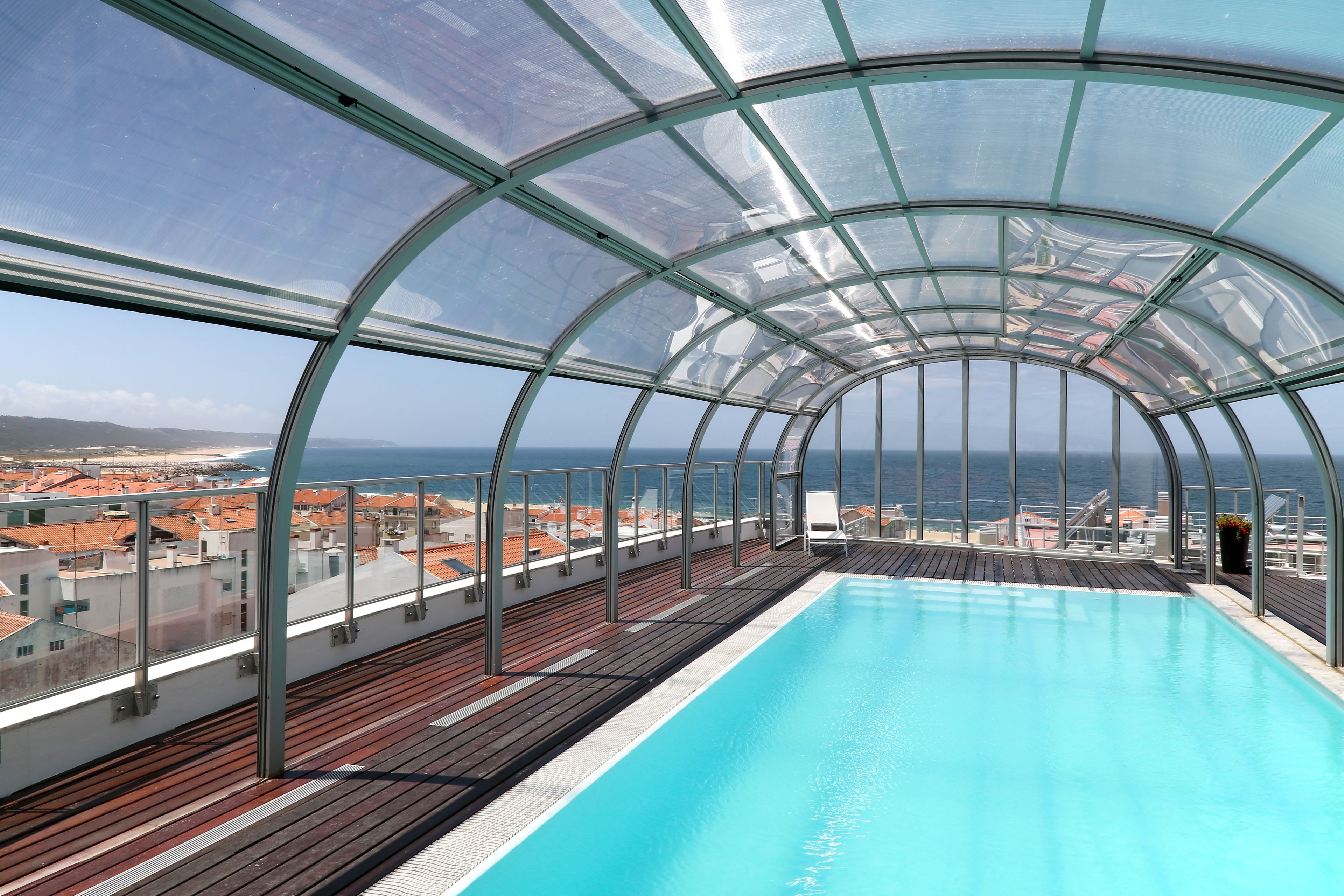A glass-enclosed infinity pool with a wooden deck, offering a view of the city and the sea.