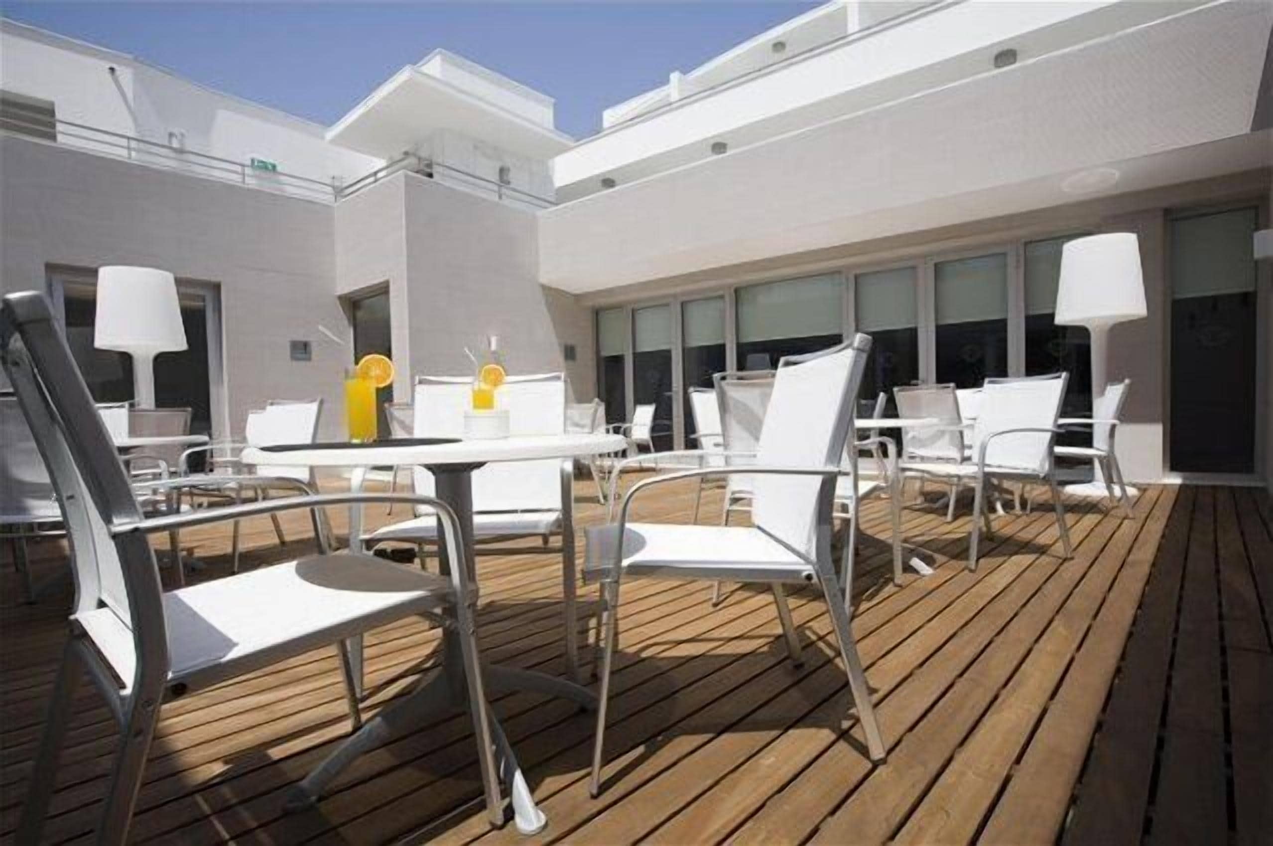 A deck with white chairs and tables, a wooden floor, and a modern building with large windows.