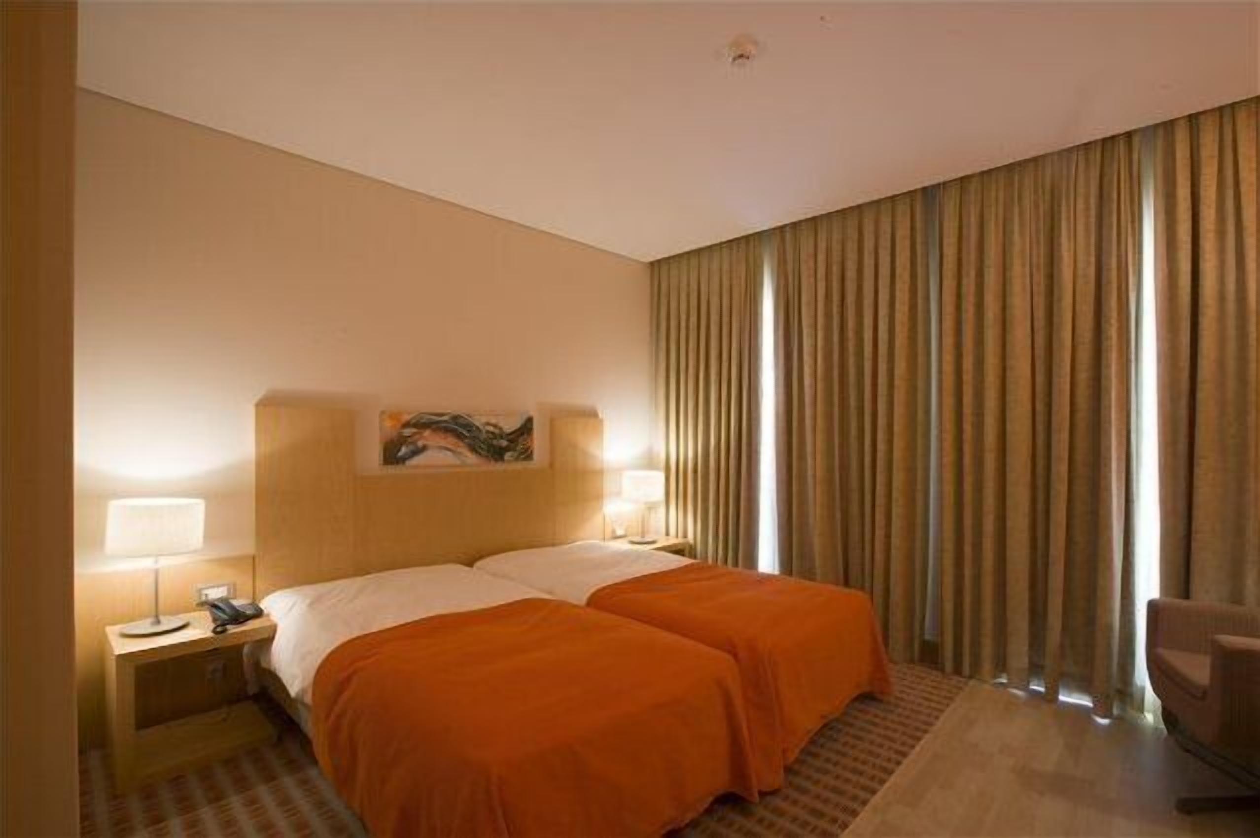Une chambre d’hôtel avec un lit double, deux tables de chevet avec des lampes, un tableau au mur et une grande fenêtre avec des rideaux.