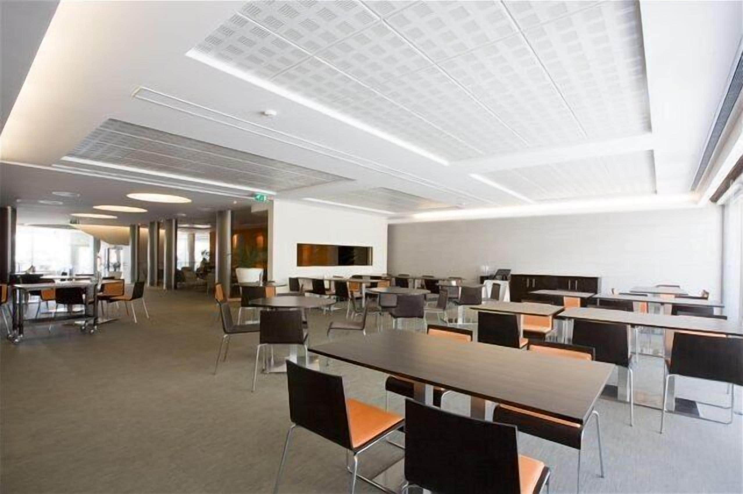 A modern conference room with multiple tables and chairs, a white ceiling with recessed lighting, and a large window providing natural light.