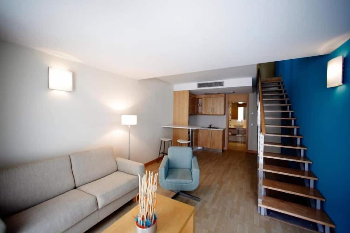 Ein modernes Wohnzimmer mit einer Couch, einem Couchtisch und einem Sessel. Eine Treppe führt zu einer Küchenzeile mit Holzschränken und einem Esstisch.