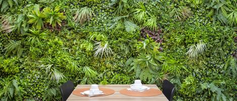 Mesa de comedor dispuesta con platos y tazas blancos, rodeada por un exuberante muro verde de diversas plantas.