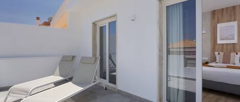 Een balkon met twee witte lounge fauteuils, een schuifdeur naar een slaapkamer met een bed en een houten hoofdbord, en een helderblauwe lucht op de achtergrond.