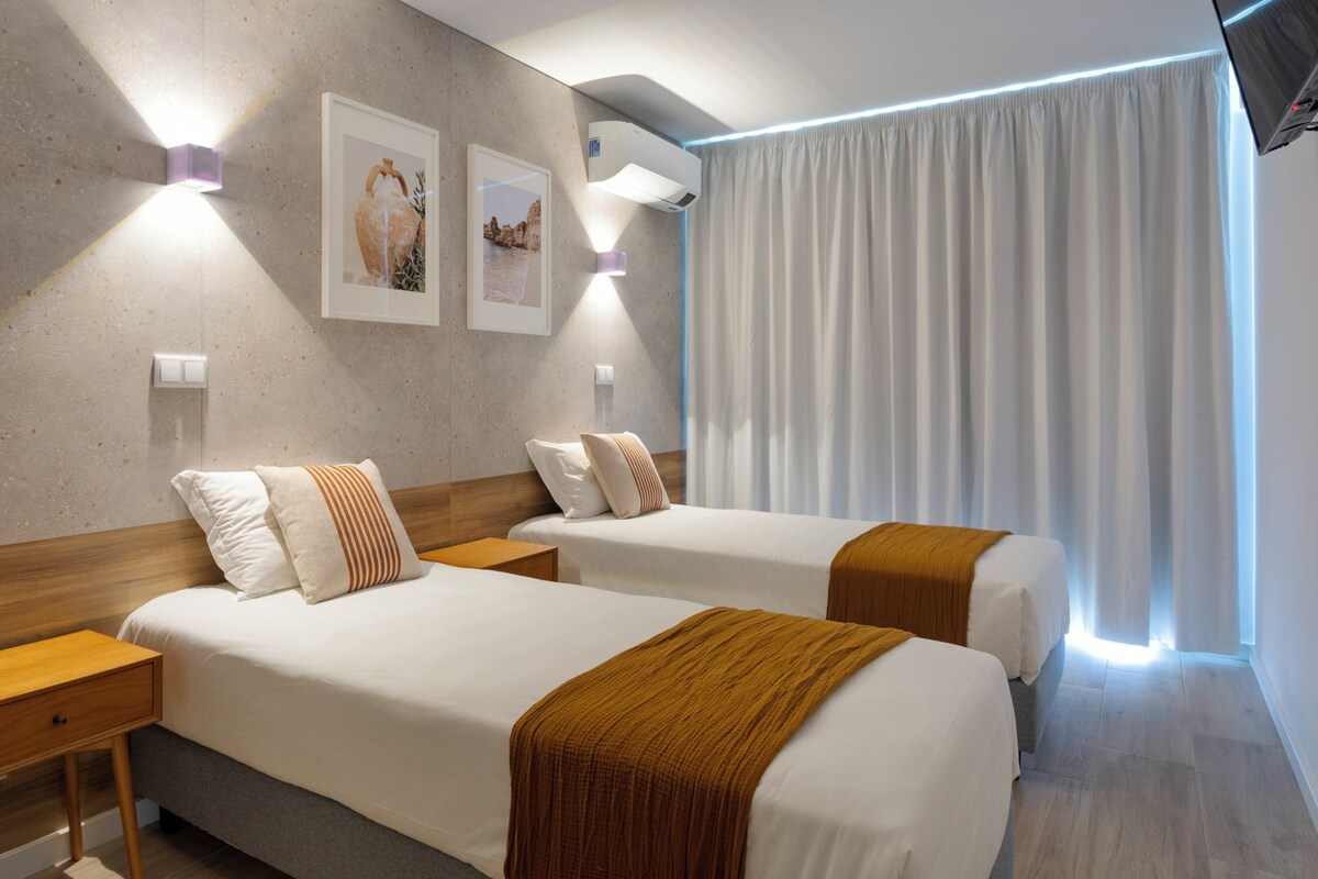 Une chambre d’hôtel avec deux lits, une table de chevet en bois, un climatiseur fixé au mur et une fenêtre avec des rideaux blancs.