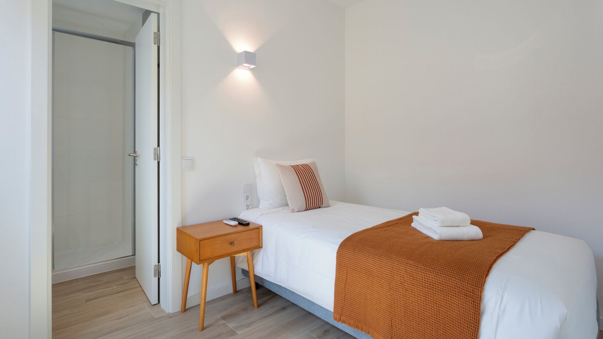 A bedroom with a bed, a nightstand, a remote control, and a wall-mounted light.