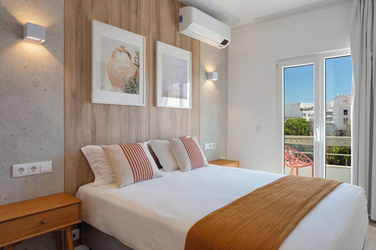 Une chambre moderne avec un grand lit, une tête de lit en bois, deux lampes fixées au mur et une vue sur la ville grâce à une porte-fenêtre coulissante.
