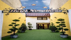 Hotel Novotheos con una entrada central, árboles topiarios en macetas y un cielo despejado.