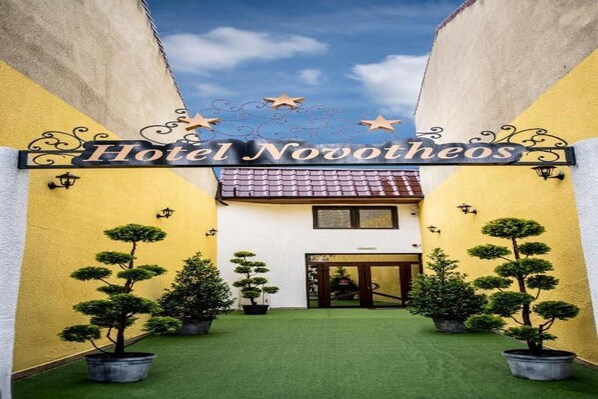 Hotel Novotheos amb una entrada central, arbres de topiaris en testos i un cel clar.