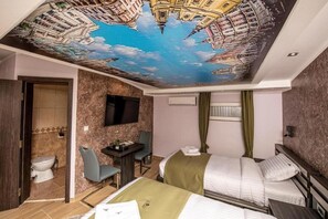 Habitació d'hotel amb un mural al sostre, un llit, una taula de treball amb cadira, un televisor i una finestra amb cortines.